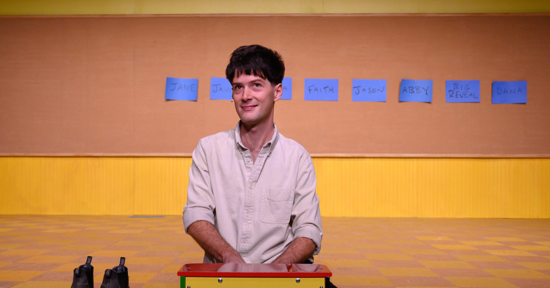 Meet the Playwright: Milo Cramer – Playwright Milo Cramer seated on a tile floor in what appears to be a classroom.