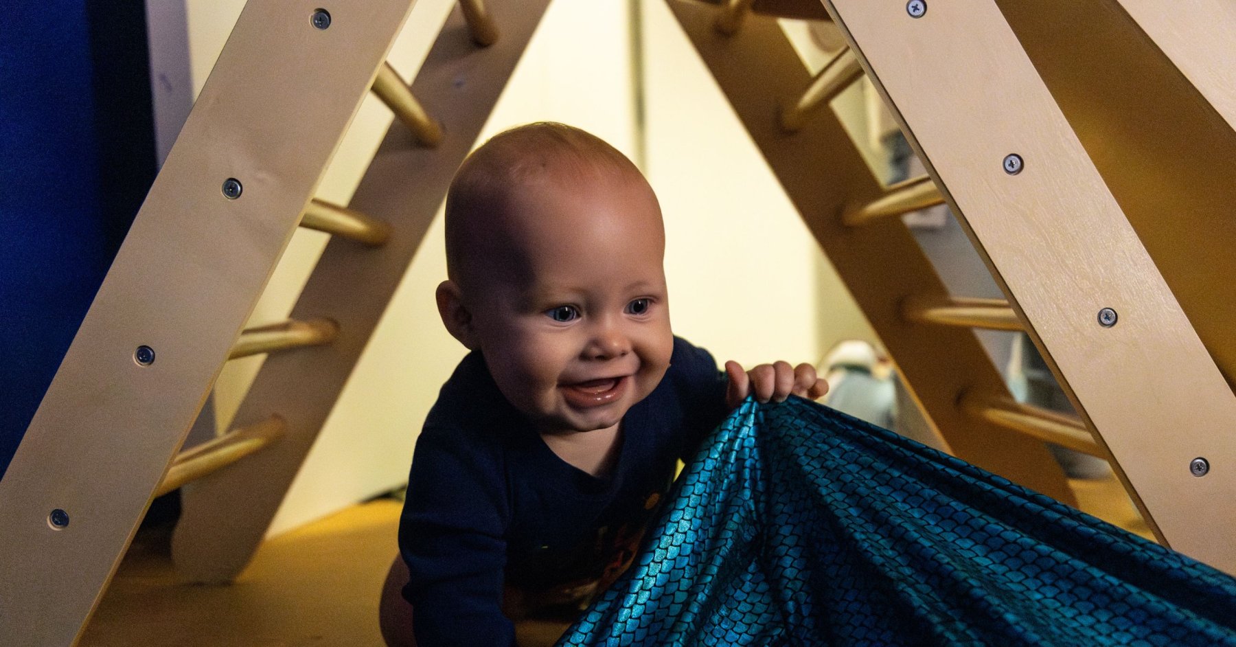 baby crawling under wooden triangle holding shiny blue fabric